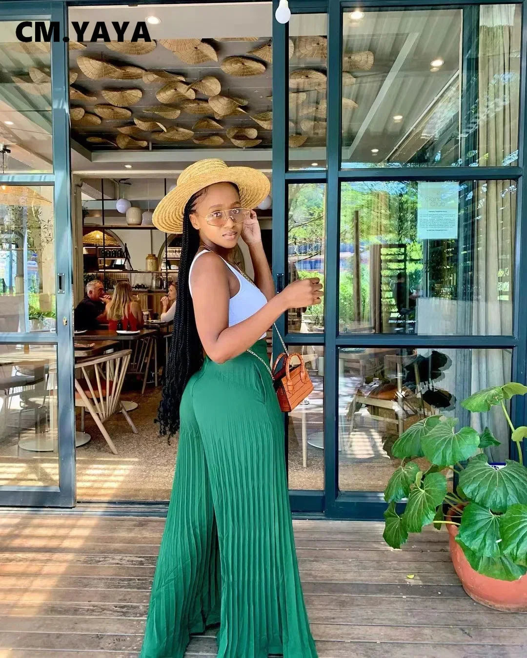 Stylish woman in straw hat, green pleated pants, white top outside modern cafe