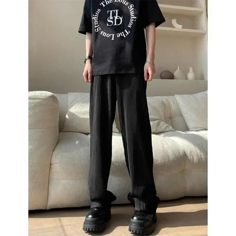 Person in black graphic t-shirt, pleated black pants, chunky shoes, standing by cream sofa