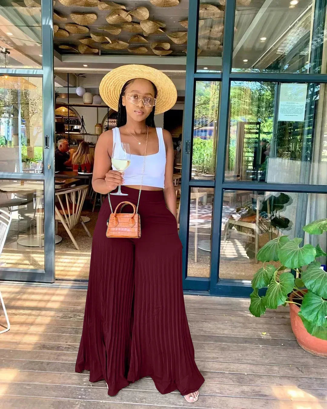 Woman in wide maroon pants, crop top, straw hat, and sunglasses holds wine outside cafe