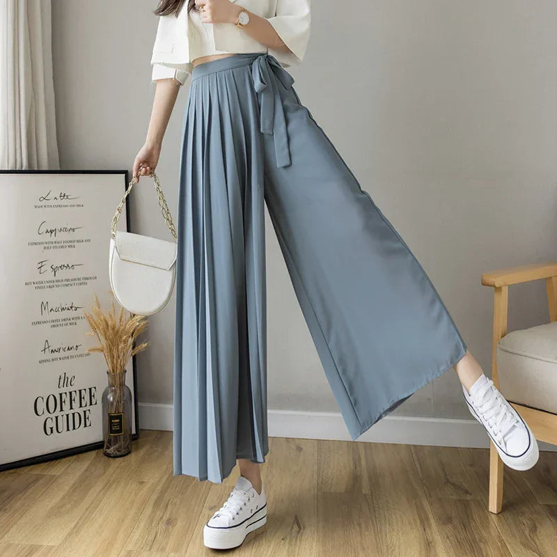 Woman in wide leg blue pleated pants, white crop top, white sneakers, and holding a handbag.