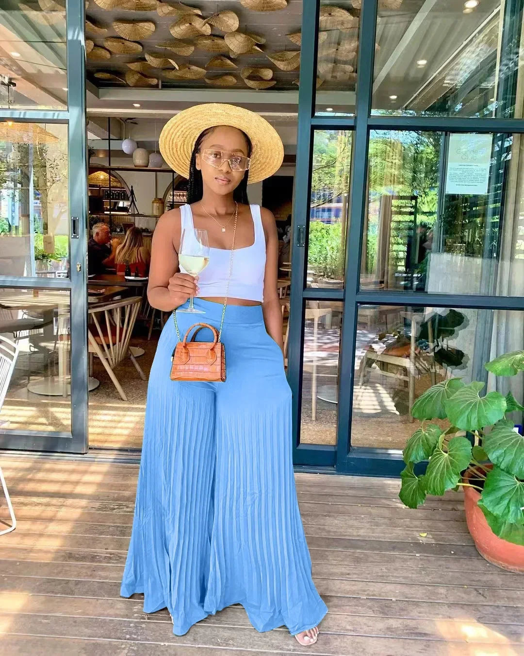 Woman in summer outfit with straw hat and blue pleated pants holding wine glass outside café