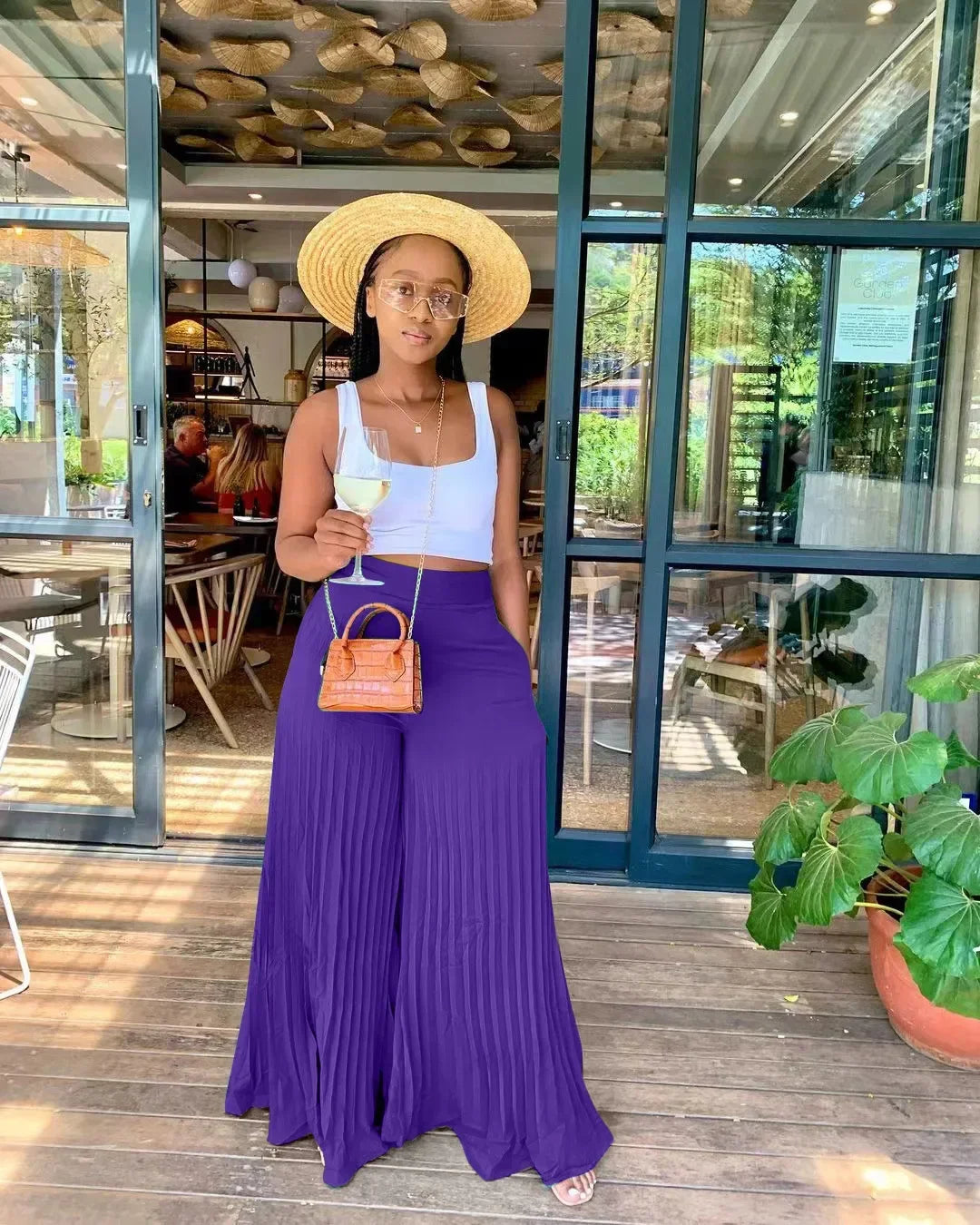 Woman in straw hat, white crop top, purple wide-leg pants, holding wine outside a restaurant