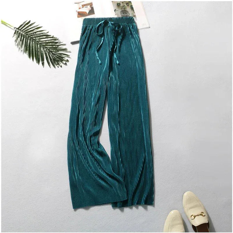 Teal pleated wide-leg pants with drawstring waist, flat-lay with loafers and palm leaf.