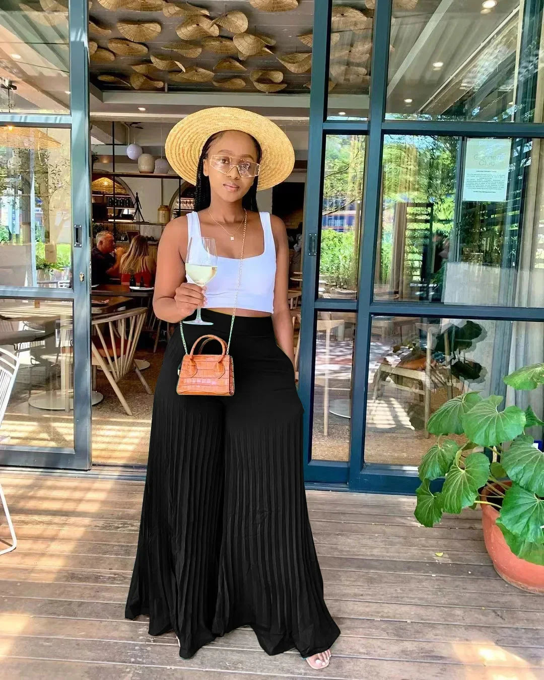 Woman in a white crop top, black pleated pants, and straw hat holding a wine glass outside a cafe