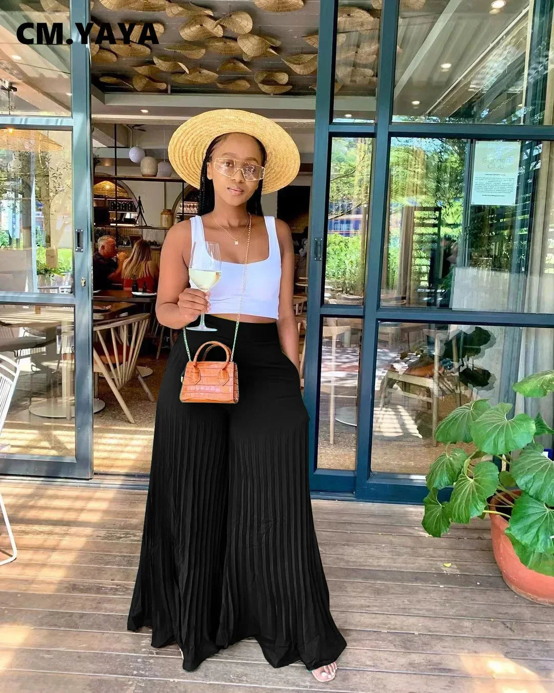 Woman in straw hat and sunglasses, wearing crop top and black wide-leg pants, holding wine