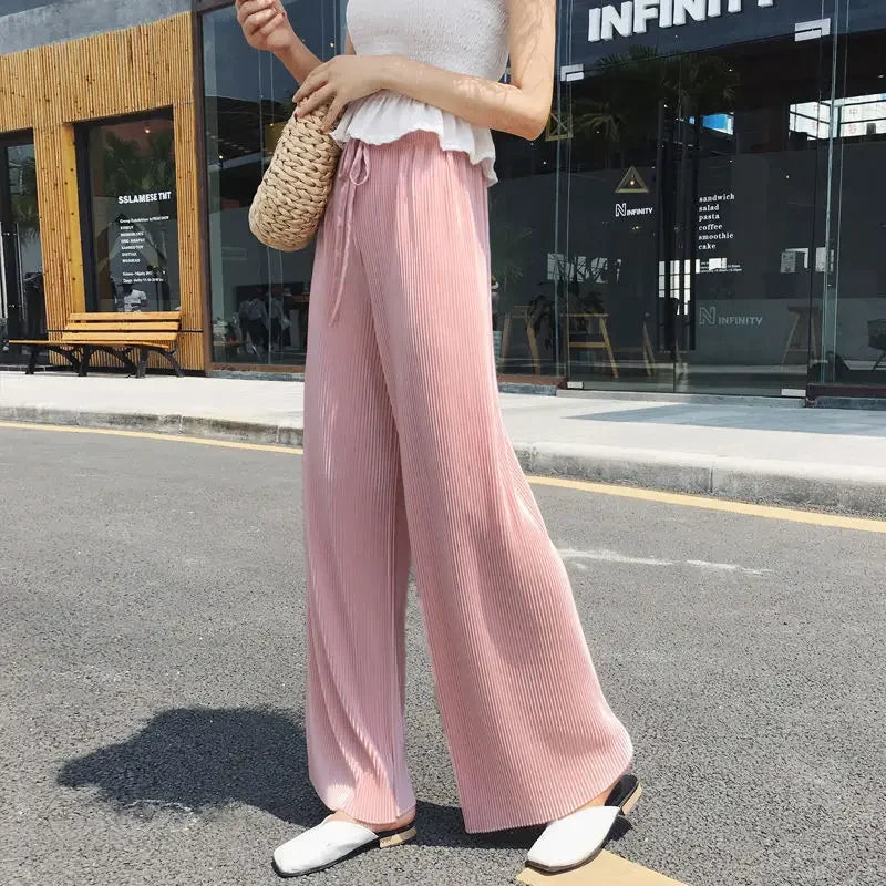 Woman wearing pink wide-leg pleated pants, white top, and holding woven bag outdoors