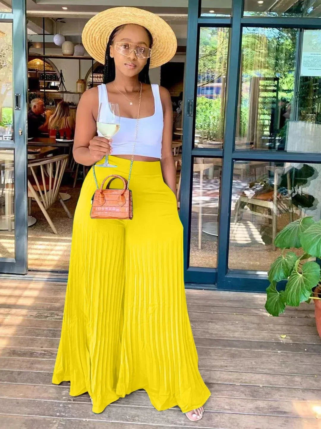 Woman in straw hat, white crop top, yellow wide-leg pants, holding wine outside cafe